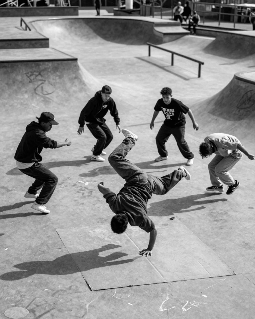A group of breakdancers spins on a cardboard mat in a sunlit skate park.