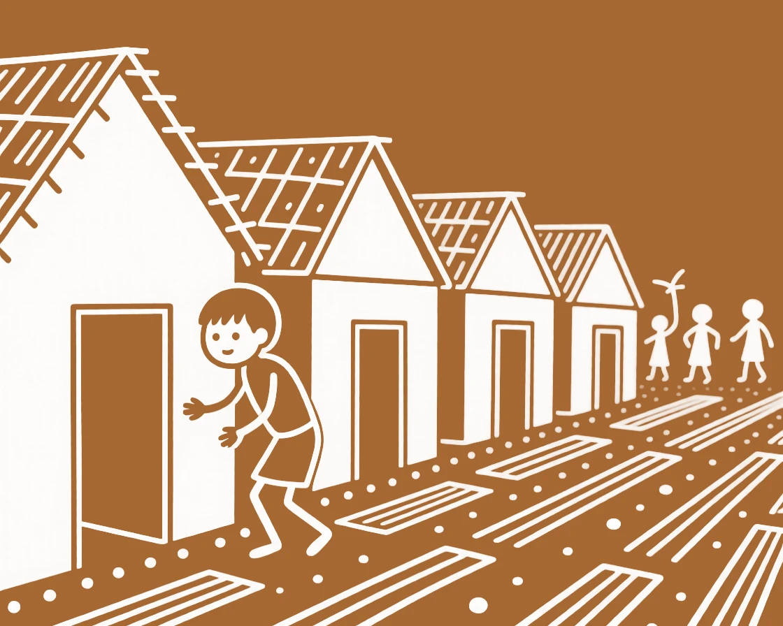 A shy young boy playing hide and seek behind a row of huts.
