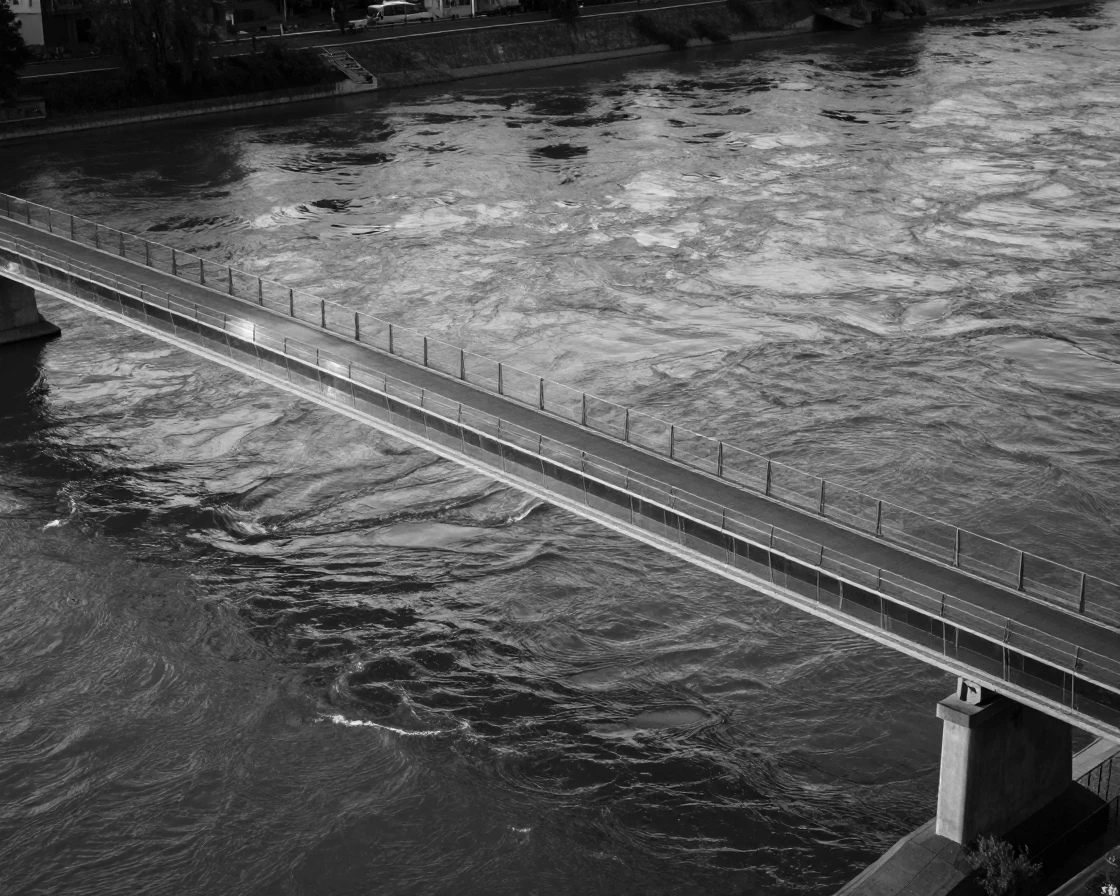 A modern glass bridge spans a wide river below beside a wide flowing river.