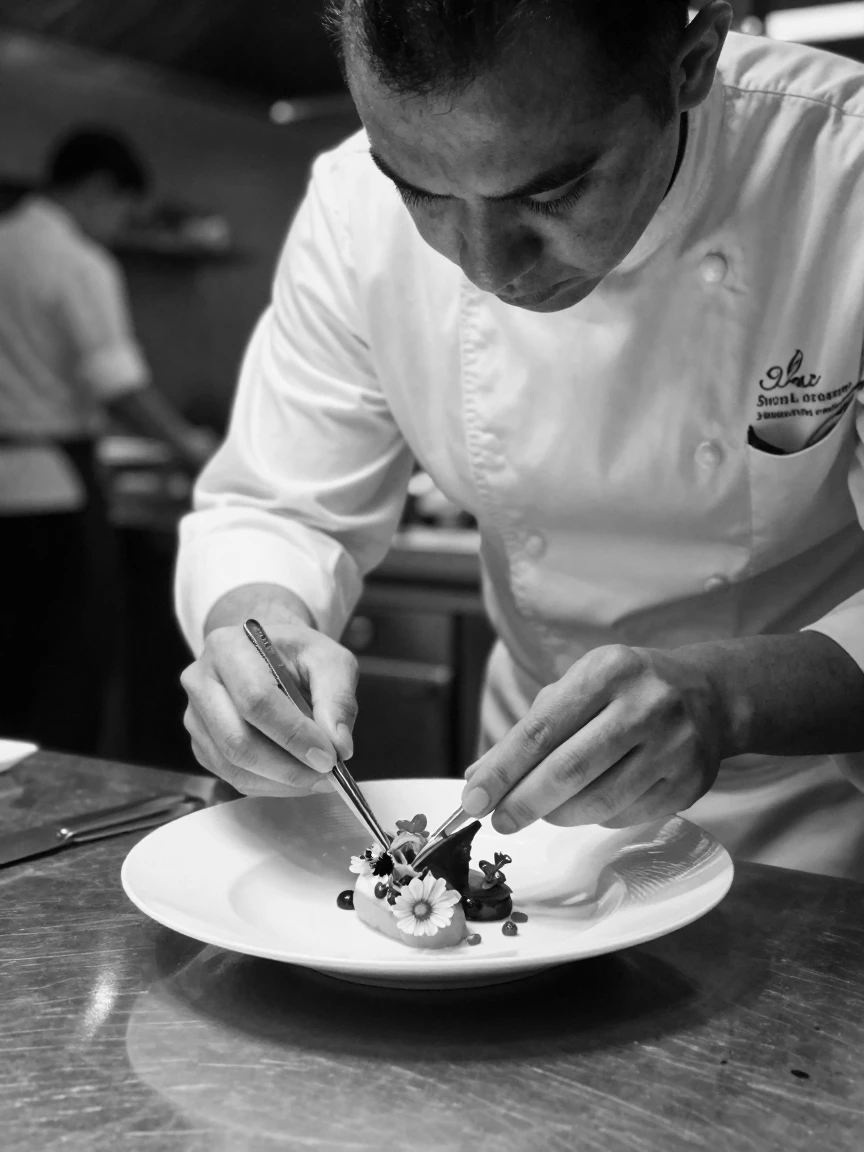 A chef plates a delicate dessert with tweezers at a fine dining pass station.